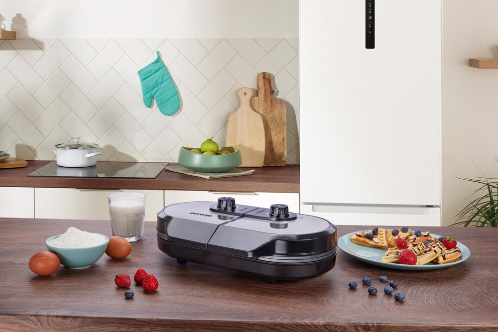 Gofrownica Gorenje na stole obok talerza z goframi, miski z mąką, jajek i truskawek