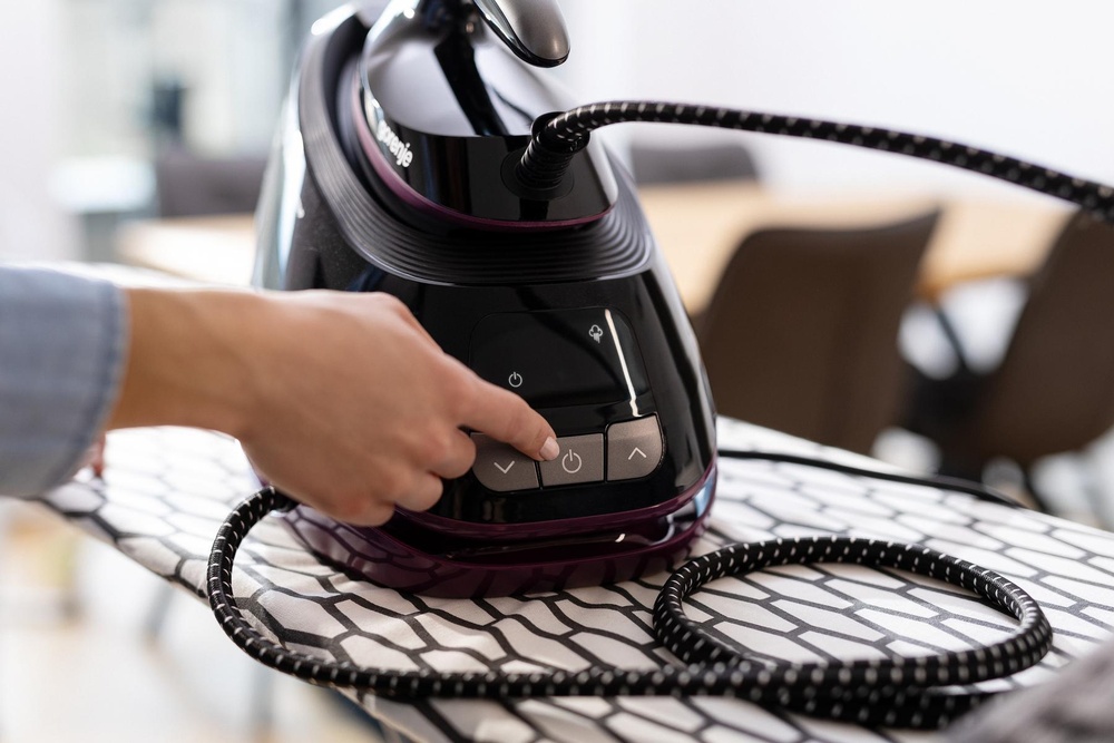Mână care apasă butonul de pornire pe panoul de control al generatorului de abur Gorenje pe masa de călcat.