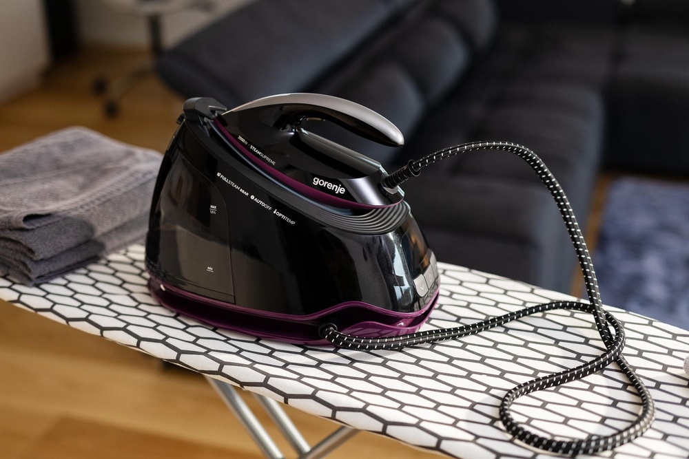 Generator de abur Gorenje negru-violet pe masa de călcat lângă un prosop gri pliat.